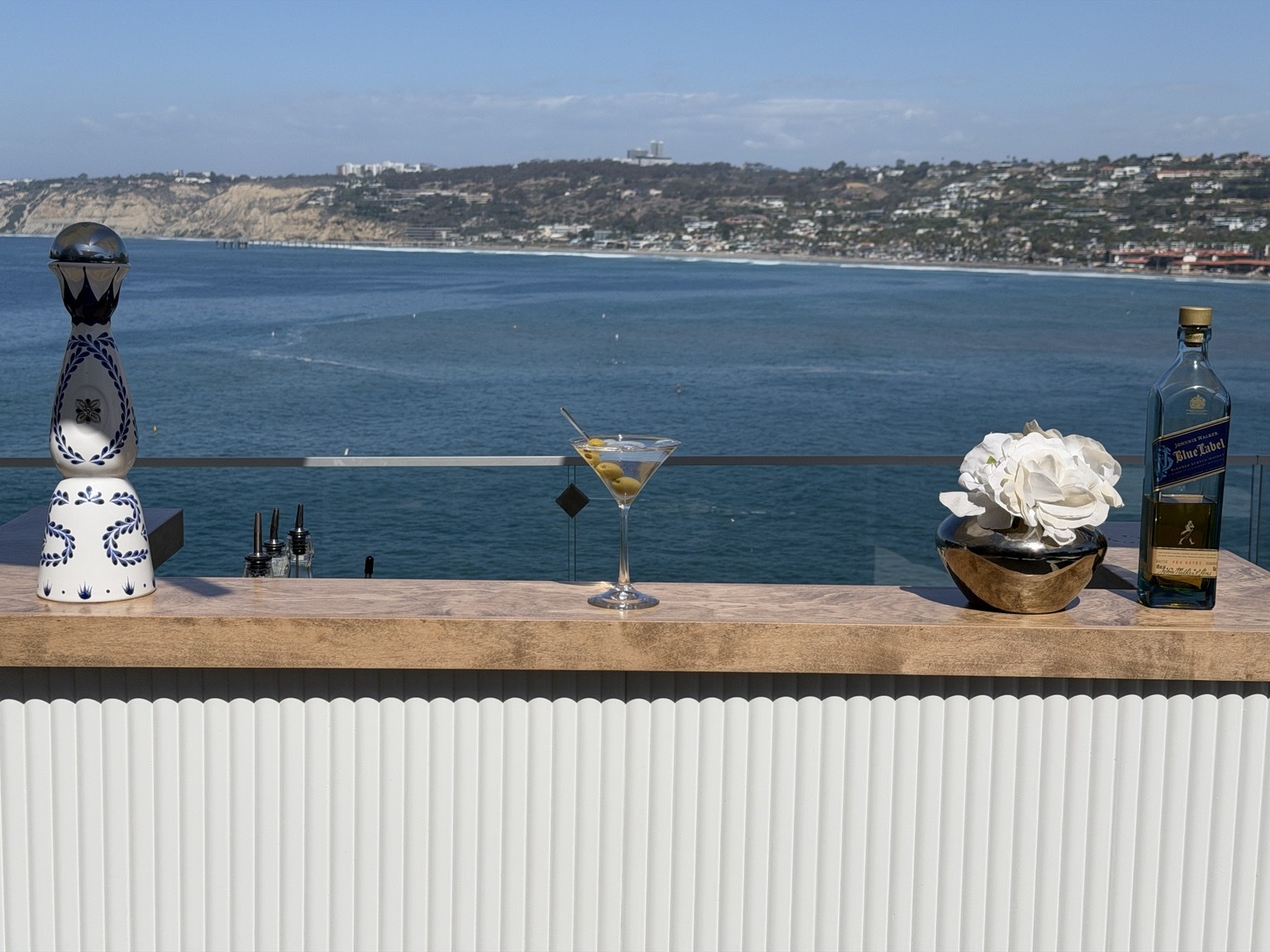 A cocktail, white flowers and bottles styled on the wood counter with an ocean backdrop